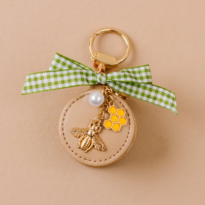 Travel Jewelry Case Bag Charm, A tan round zip case charm with a green gingham bow, a pearl, a gold bee, and a honeycomb charm, in the Hey Honey design.