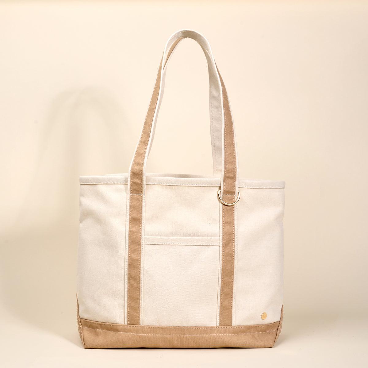 The Signature Canvas Tote Bag, A natural canvas tote bag with black webbing straps, a front pocket, and a gold D-ring, shown against a cream background.