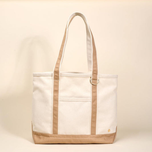 The Signature Canvas Tote Bag, A natural canvas tote bag with black webbing straps, a front pocket, and a gold D-ring, shown against a cream background.