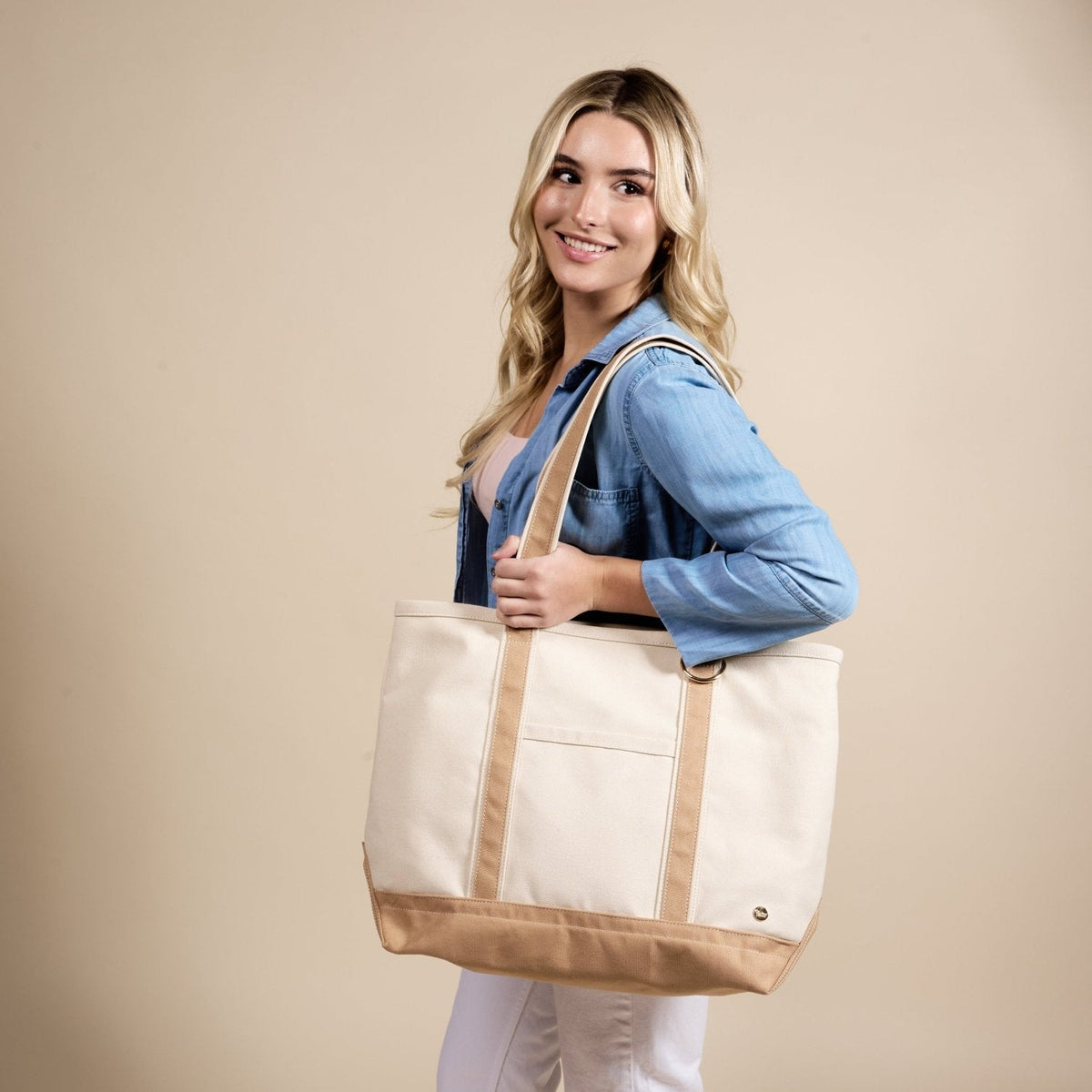 The Signature Canvas Tote Bag, A natural canvas tote bag with black webbing straps, a front pocket, and a gold D-ring, shown against a cream background.
