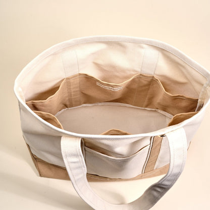 The Signature Canvas Tote Bag, A natural canvas tote bag with black webbing straps, a front pocket, and a gold D-ring, shown against a cream background.