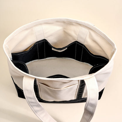 The Signature Canvas Tote Bag, A natural canvas tote bag with black webbing straps, a front pocket, and a gold D-ring, shown against a cream background.