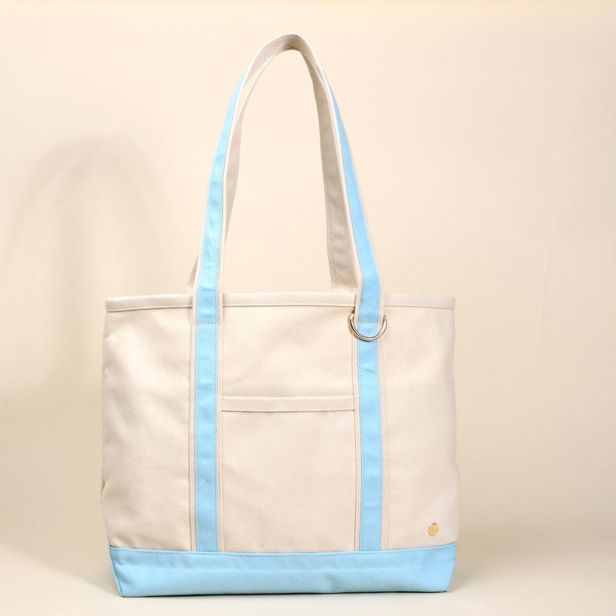 The Signature Canvas Tote Bag, A natural canvas tote bag with black webbing straps, a front pocket, and a gold D-ring, shown against a cream background.