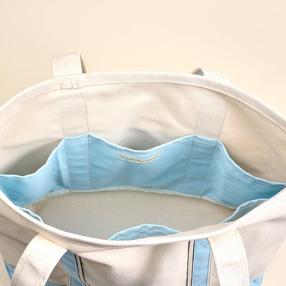 The Signature Canvas Tote Bag, A natural canvas tote bag with black webbing straps, a front pocket, and a gold D-ring, shown against a cream background.
