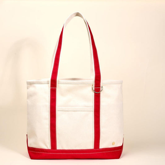 The Signature Canvas Tote Bag, A natural canvas tote bag with black webbing straps, a front pocket, and a gold D-ring, shown against a cream background.