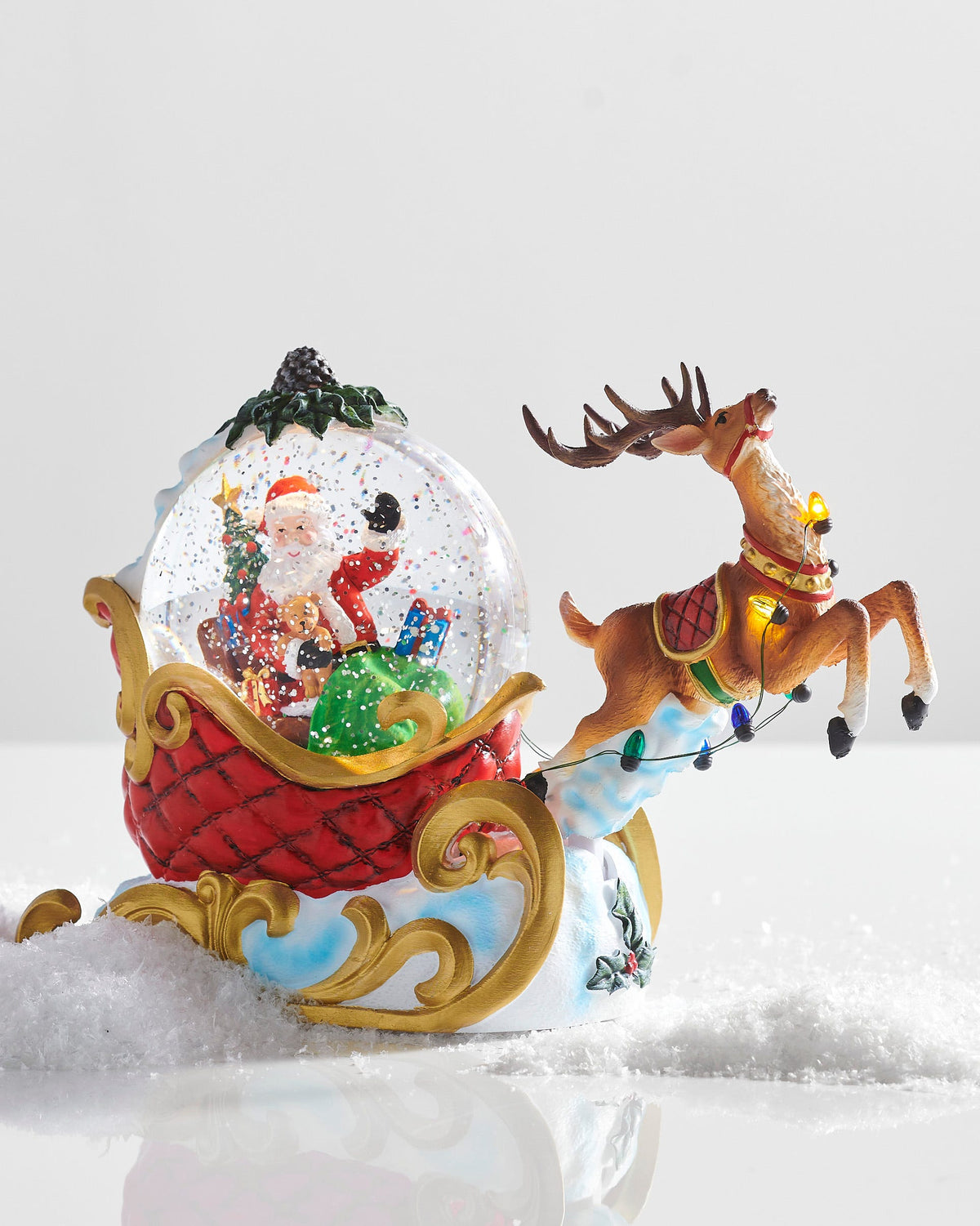 Animated Santa's Sleigh and Reindeer Lighted Water Globe, Santa with a sack of toys in a glowing water globe that rides in a red quilted sleigh on gold scrolled runners, with a reindeer in mid-leap on a cloud ahead of it.