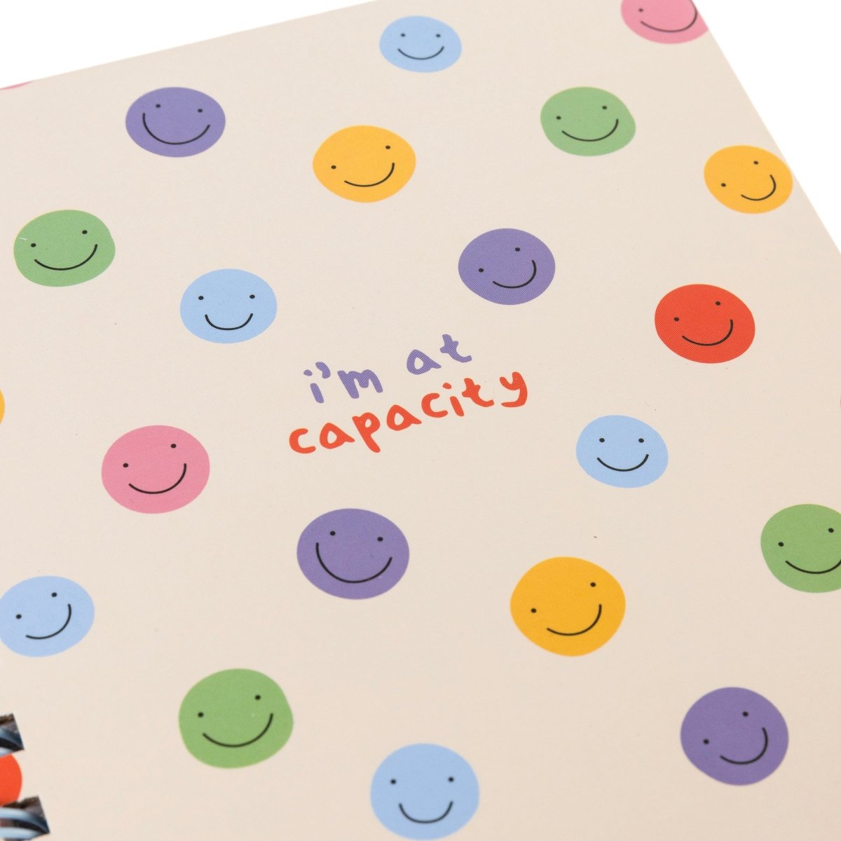 At Capacity Everyday Journal, A cream spiral-bound journal covered in rainbow smiley faces with the words i'm at capacity in colorful lettering at the center.