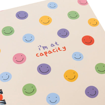 At Capacity Everyday Journal, A cream spiral-bound journal covered in rainbow smiley faces with the words i'm at capacity in colorful lettering at the center.