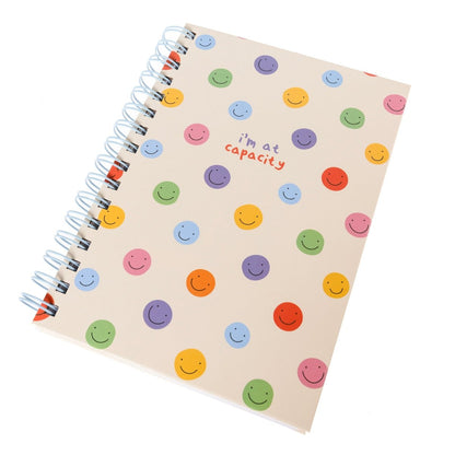 At Capacity Everyday Journal, A cream spiral-bound journal covered in rainbow smiley faces with the words i'm at capacity in colorful lettering at the center.
