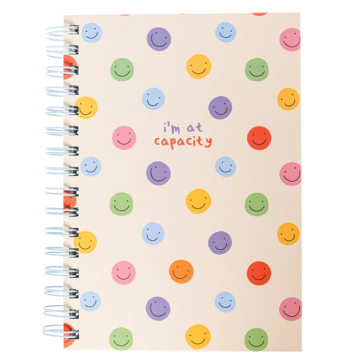 At Capacity Everyday Journal, A cream spiral-bound journal covered in rainbow smiley faces with the words i'm at capacity in colorful lettering at the center.