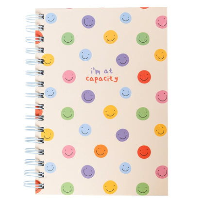 At Capacity Everyday Journal, A cream spiral-bound journal covered in rainbow smiley faces with the words i'm at capacity in colorful lettering at the center.