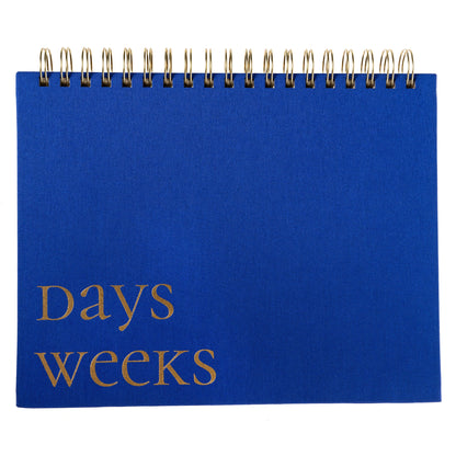 Cobalt Blue Fabric-Bound Weekly Desk Pad, A cobalt blue fabric desk pad reading days weeks in gold foil with a spiral top binding.