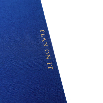 Cobalt Blue Fabric-Bound Task Journal, A cobalt blue fabric-covered task journal with gold twin wire spirals and two navy ribbon bookmark tabs, closed with an elastic band.