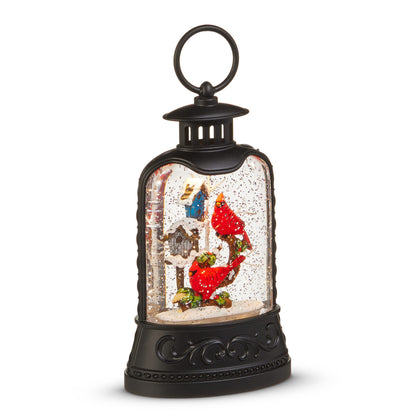 Cardinal and Birdhouse Lighted Water Lantern, A matte black lantern with a ring handle holding two red cardinals on a holly branch beside a blue birdhouse and a gray birdhouse, surrounded by swirling glitter.