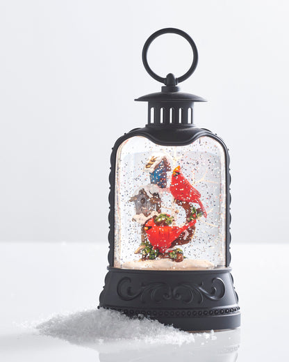 Cardinal and Birdhouse Lighted Water Lantern, A matte black lantern with a ring handle holding two red cardinals on a holly branch beside a blue birdhouse and a gray birdhouse, surrounded by swirling glitter.