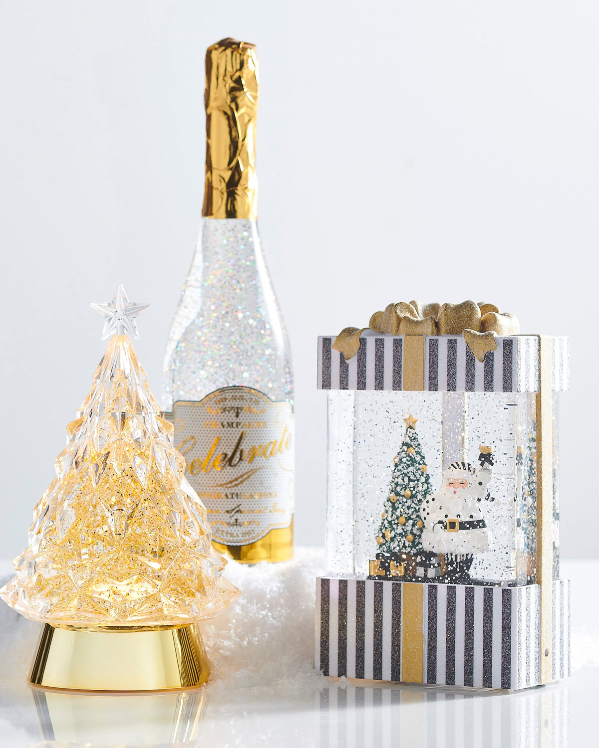 Champagne Service Santa Lighted Water Present, A black-and-white striped present-shaped lantern with a gold glitter bow, showing Santa in a white polka dot suit raising a champagne glass beside a flocked tree with gold ornaments.