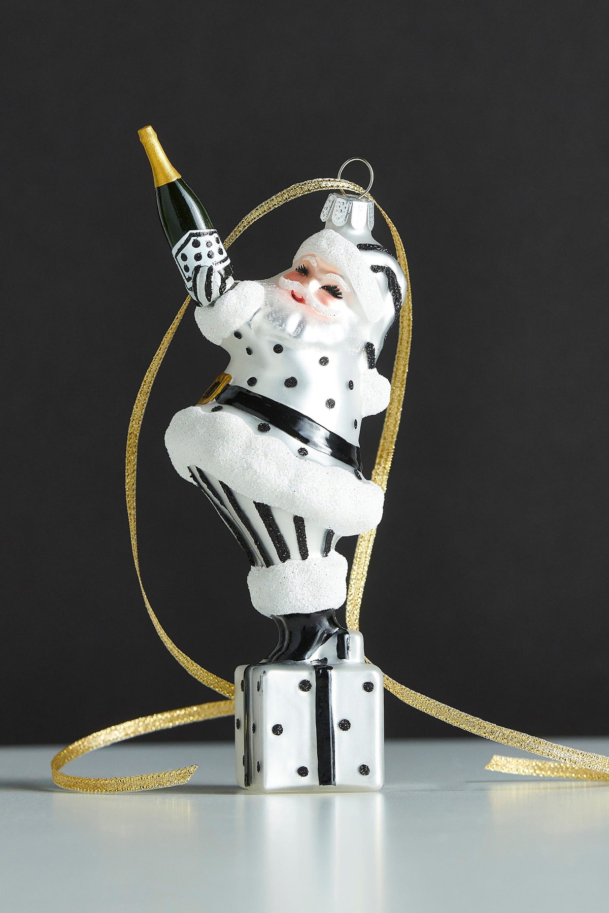 Champagne Popping Santa Ornament, A glass and resin Santa ornament in black and white polka dots, holding a champagne bottle while standing on a wrapped gift.