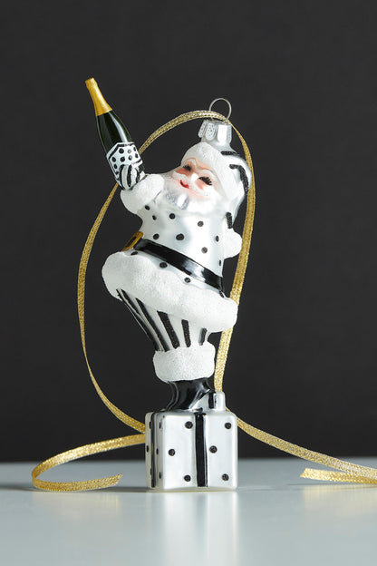 Champagne Popping Santa Ornament, A glass and resin Santa ornament in black and white polka dots, holding a champagne bottle while standing on a wrapped gift.