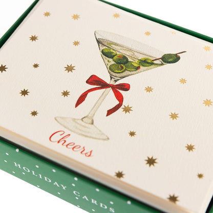 Cheers Holiday Cards, Box of 15, A cream holiday card with a martini glass, green olives and a red bow under gold foil stars and Cheers in red script, set in its green box.