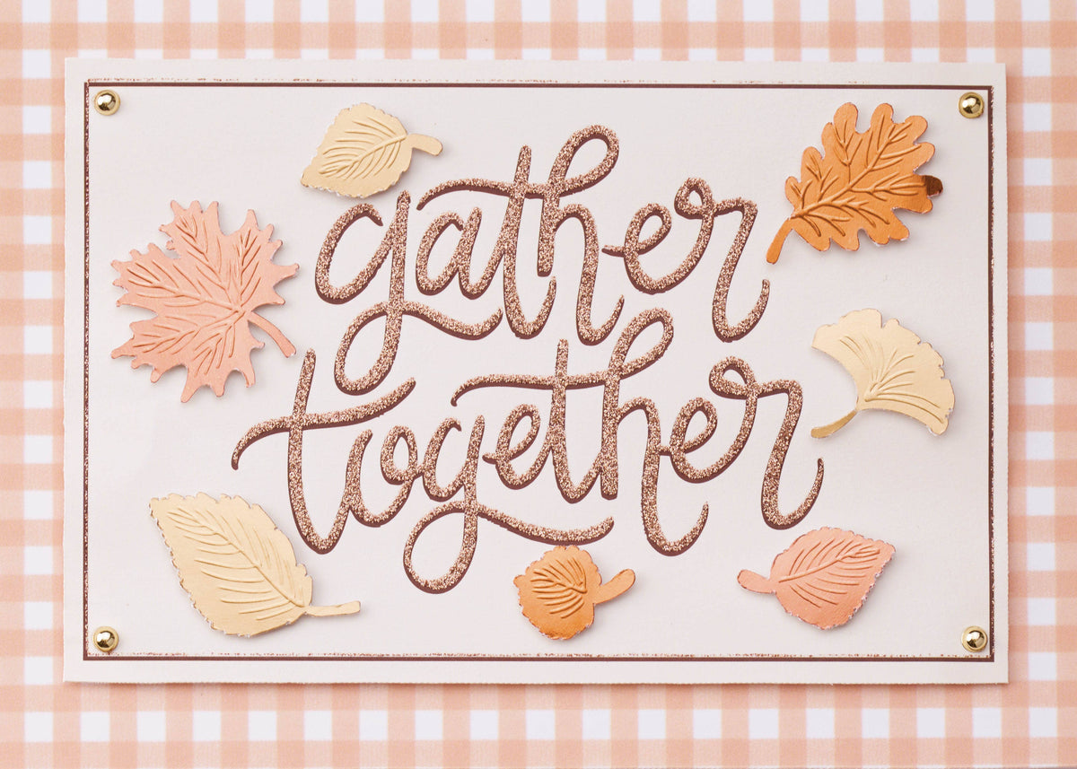 Gather in Thankfulness Greeting Card, A landscape card front with the words gather together in glittery rose-gold script on a cream panel, surrounded by blush, gold and copper leaves, with a peach gingham border and a gold stud in each corner.