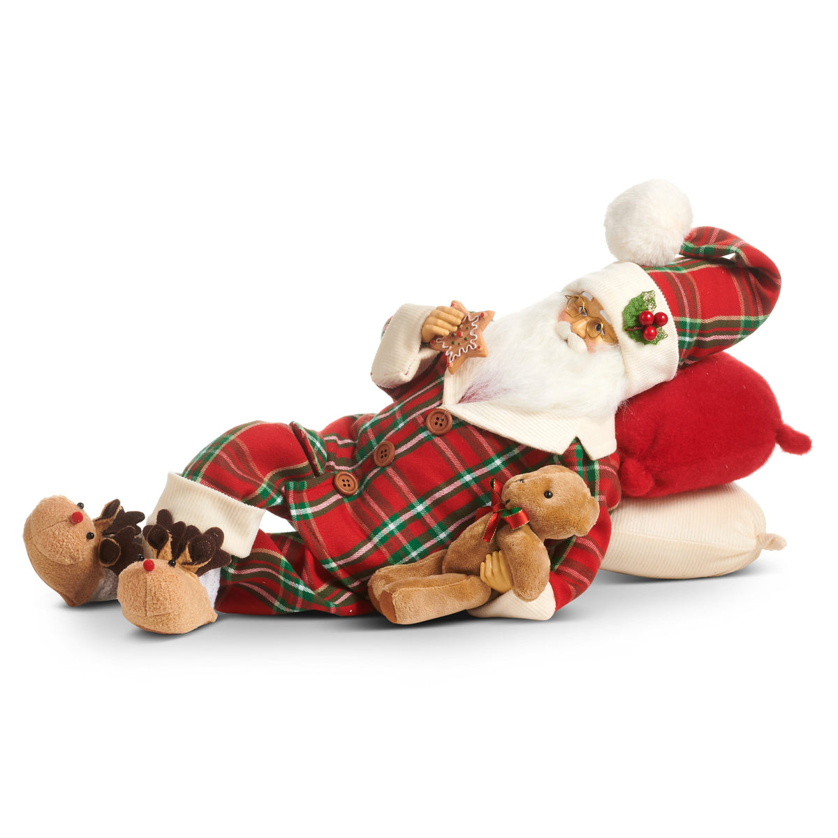 Goodnight Santa Reclining Figure, A Santa figure in red plaid pajamas and a pom-pom nightcap reclines on red and cream pillows, holding a teddy bear and an iced star cookie, with reindeer slippers on his feet.
