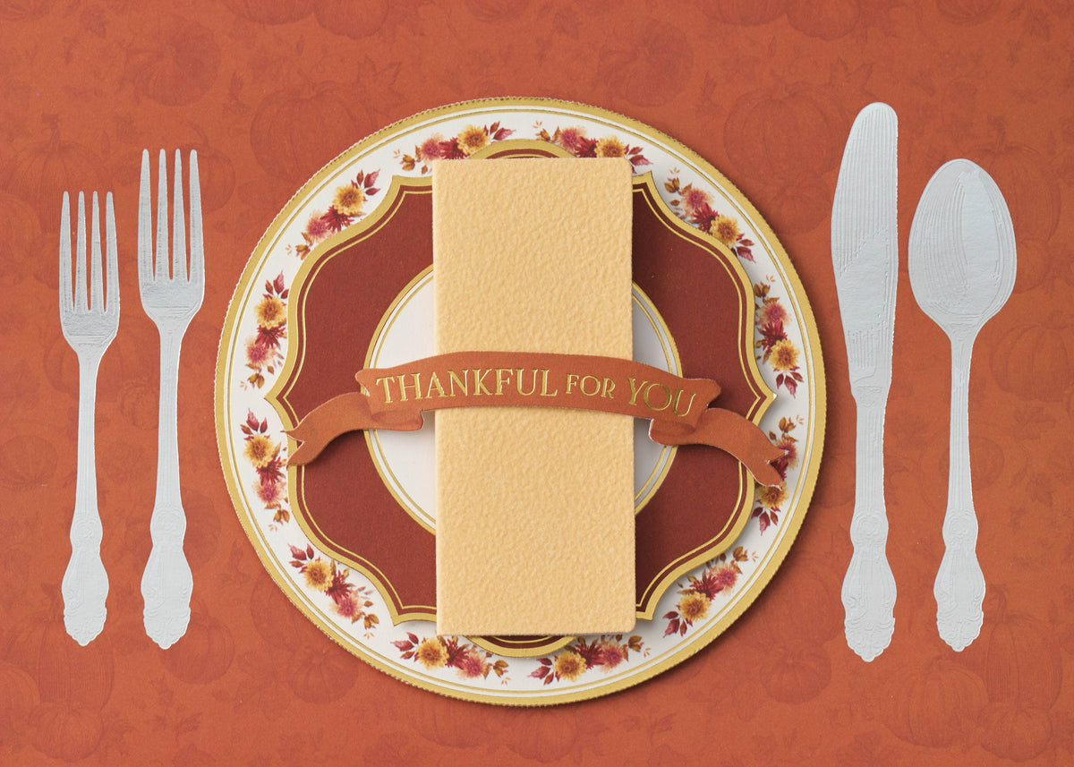 Harvest Table Greeting Card, A rust-orange card front showing a cream plate with a gold edge and floral rim, a folded napkin with a banner reading THANKFUL FOR YOU, two forks on the left and a knife and spoon on the right.
