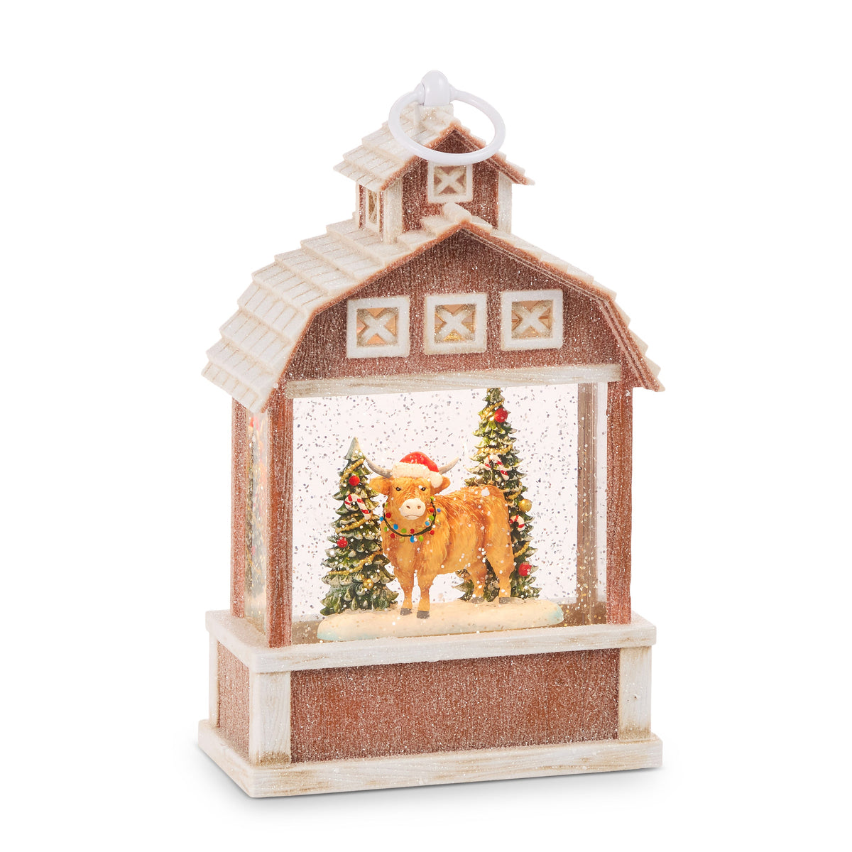 Highland Cow Lighted Water Lantern, Brown barn water lantern with a cupola and ring handle, showing a Highland cow in a Santa hat and string lights between two decorated trees.