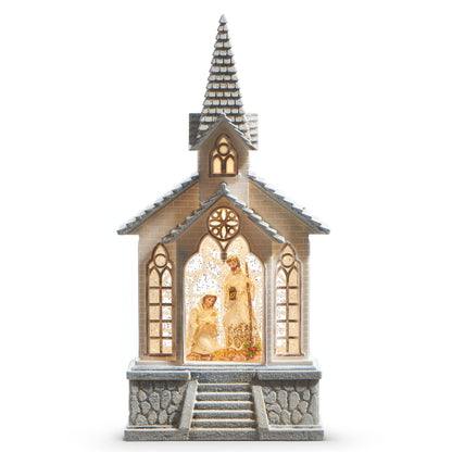 Holy Family Musical Lighted Water Church, A white church with a snow dusted shingled steeple and gray stone steps, its center arched window lit to show the Holy Family behind swirling glitter.