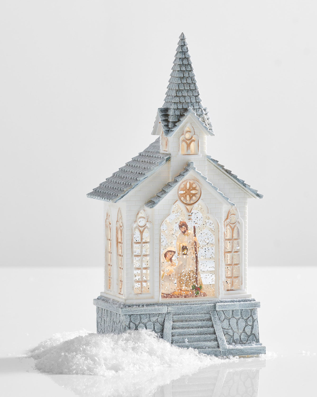 Holy Family Musical Lighted Water Church, A white church with a snow dusted shingled steeple and gray stone steps, its center arched window lit to show the Holy Family behind swirling glitter.