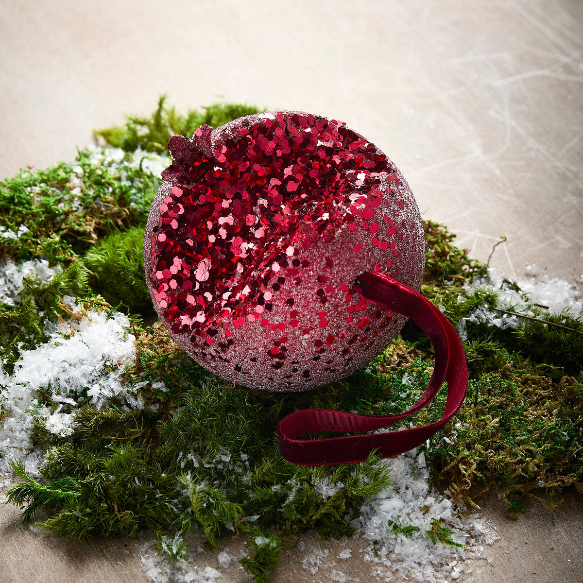 Iced Pomegranate Ornament, A red glitter ornament shaped like a pomegranate with a crowned top, hanging from a burgundy ribbon.