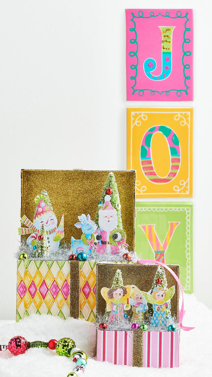 Lighted Choir Angel Vignette Box, An open pink and white striped box with a gold glitter lid, holding three paper angels, two small bottle brush trees, silver tinsel and colored baubles lit from inside.
