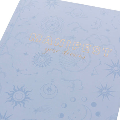Manifest Self-Care Journal, A periwinkle blue fabric journal printed with celestial suns, moons, and stars under 'Manifest Your Dreams' in gold foil.