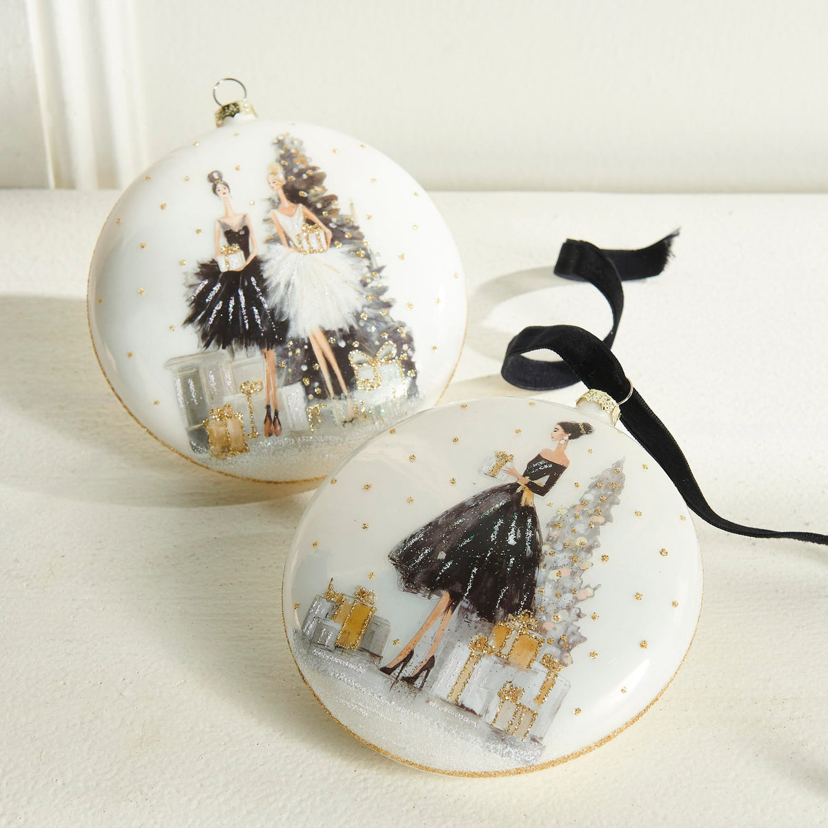 Moonlit Gala Glass Disc Ornament, Two white glass disc ornaments with gold glitter dots, one showing a woman in a black dress with gifts beside a tree and the other showing two women in black and white dresses holding gifts.