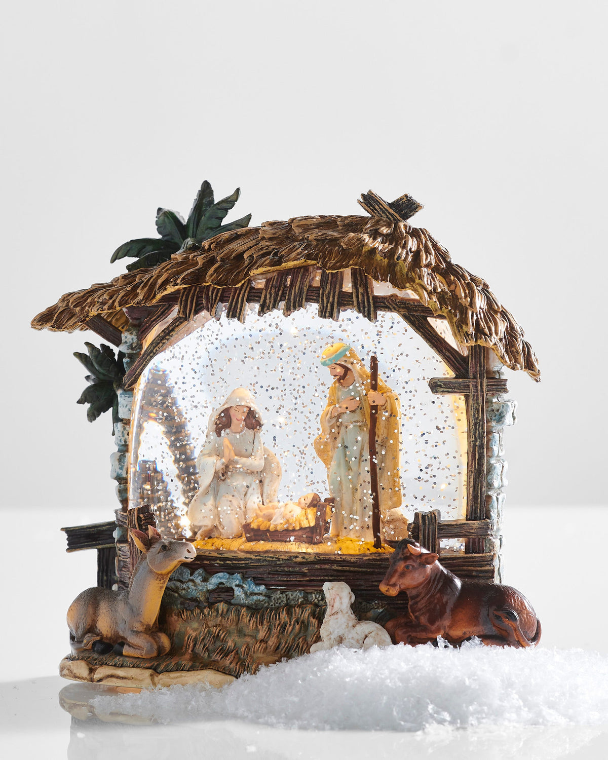 Nativity Stable Lighted Water Lantern, A resin nativity stable with a thatched roof and palms, Mary and Joseph at the manger behind glittered water, and a donkey, a lamb and an ox resting on the base.