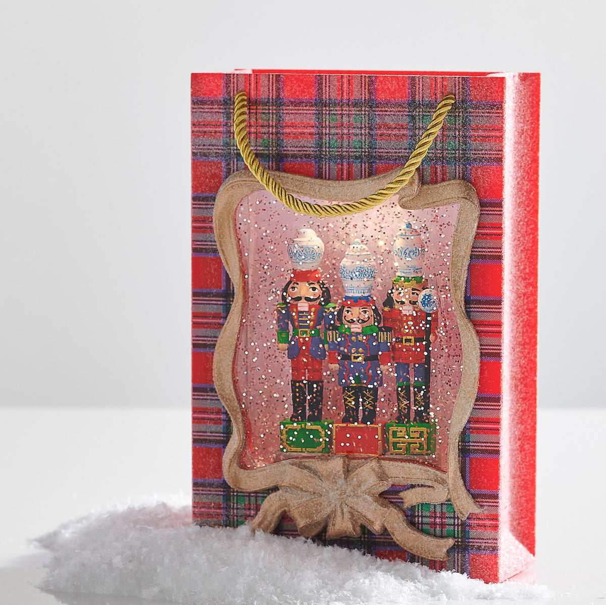 Noble Nutcracker Lighted Water Present, A red plaid gift-bag-shaped lantern with gold rope handles and a gold ribbon frame, showing three nutcrackers each topped with a blue-and-white ginger jar amid glitter.