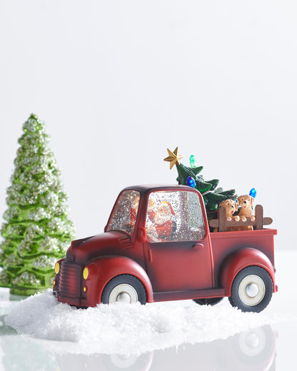 Santa and Pups Lighted Water Truck, A red vintage-style pickup truck with Santa waving inside a glitter-filled cab and two tan pups looking over a wooden rail in the bed beside a small Christmas tree lit with colored bulbs.