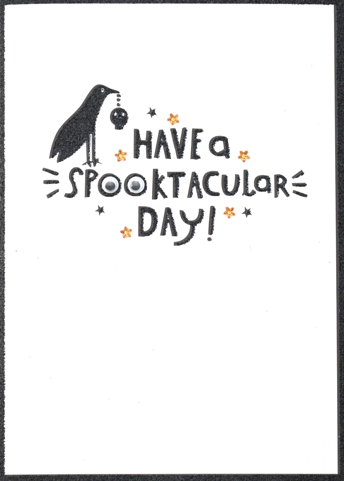 Spooktacular Wishes Greeting Card, Front of the card showing a black crow holding a tiny skull above the words HAVE a SPOOKTACULAR DAY! with a pair of googly eyes in the double O and orange star-shaped gems on a white ground.