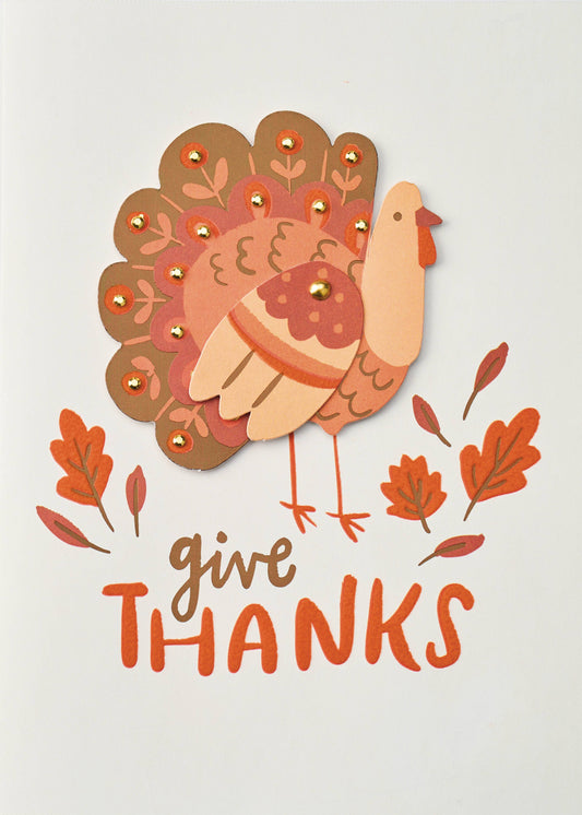Thanksful Little Turkey Greeting Card, An off-white card front with a layered turkey in peach, coral and bronze, its tail set with small gold gems, above oak leaves and the words give THANKS.