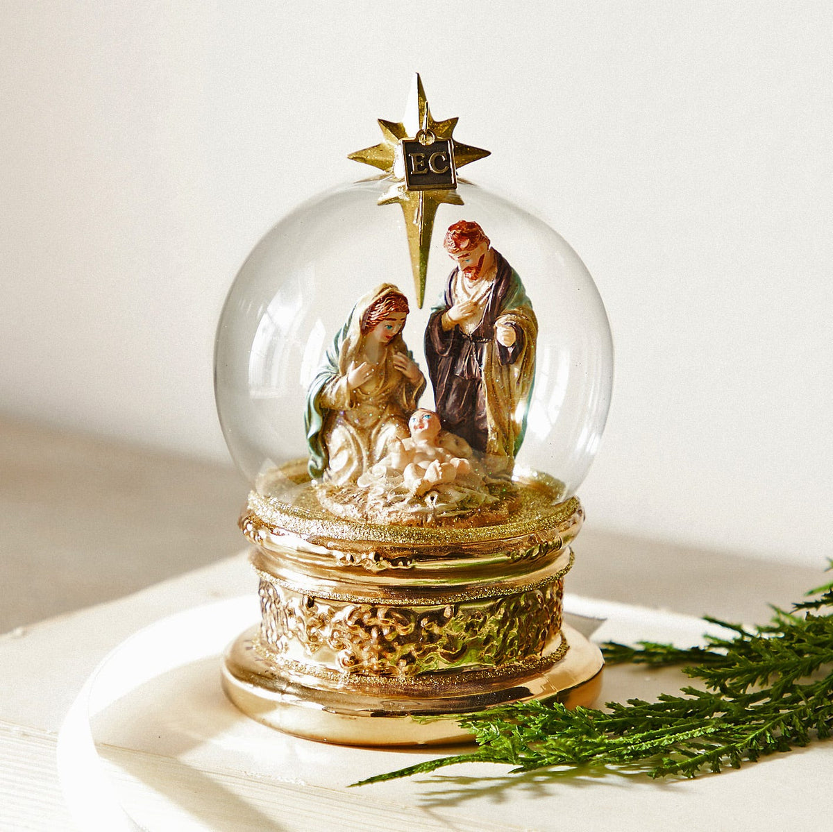 The Blessed Birth Globe Ornament, A clear glass globe ornament with a nativity scene of Mary, Joseph and the baby, set on a gold toned base.