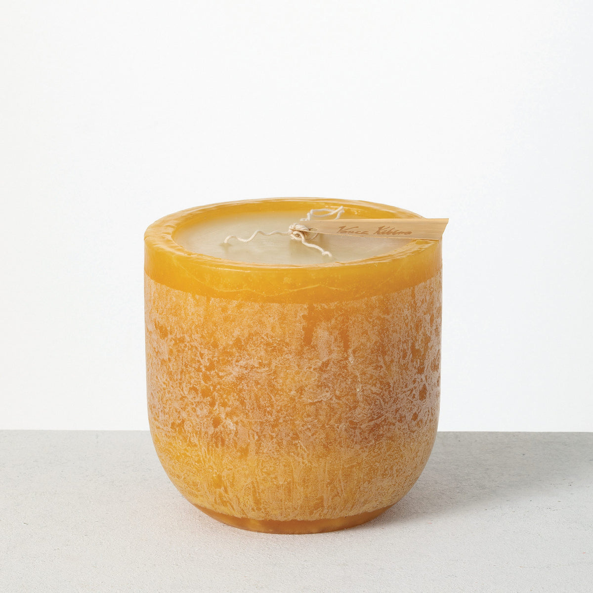 Timber Goblet Candle 6 x 6 Inch, A round, cup-shaped candle in golden amber wax with a mottled, bark-like surface, a wide cream wax top holding a looped wick, and a wooden tag reading Vance Kitira resting across the rim.