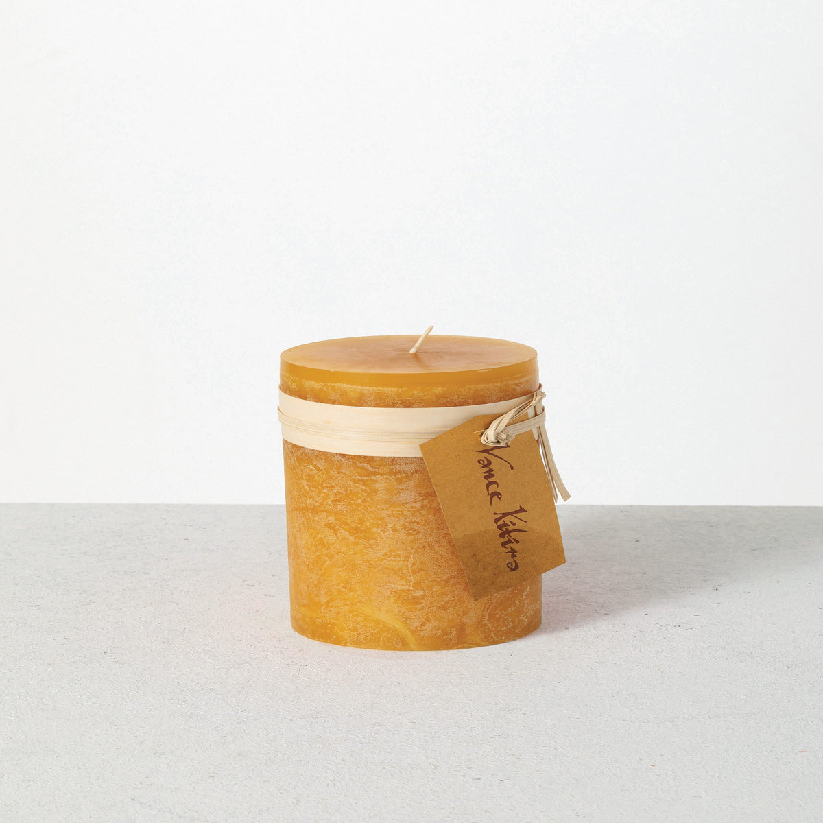 Timber Pillar Candle 3.25 x 3 Inch, A short pillar candle in warm honey gold sits on a plain background with a cream band and a paper tag around its middle.