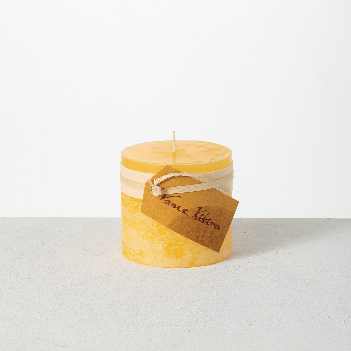 Timber Pillar Candle 3.25 x 3 Inch, A short pillar candle in warm honey gold sits on a plain background with a cream band and a paper tag around its middle.