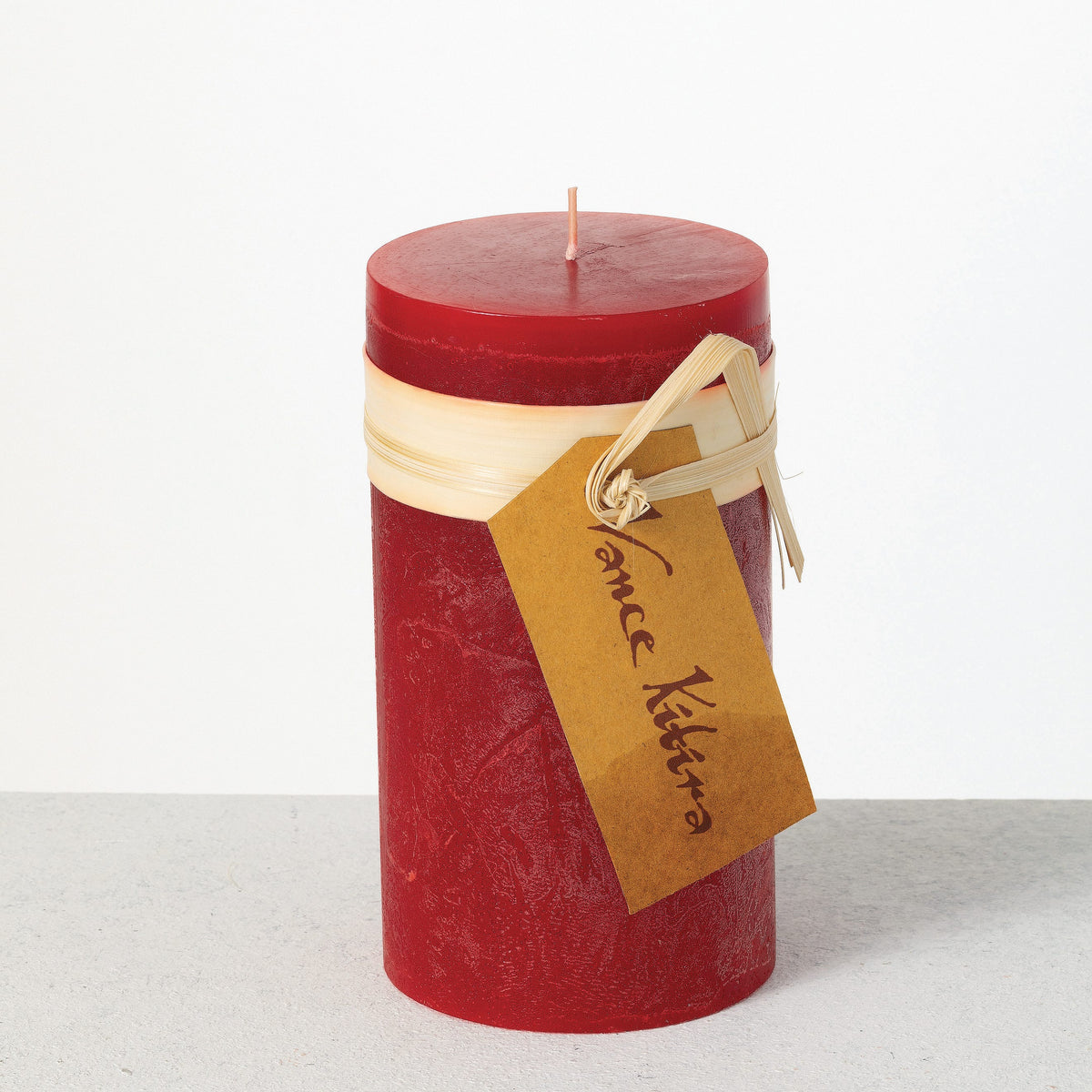 Timber Pillar Candle 3.25 x 6 Inch, A tall reddish brown pillar candle with a textured surface stands tied with a paper band and a hang tag.