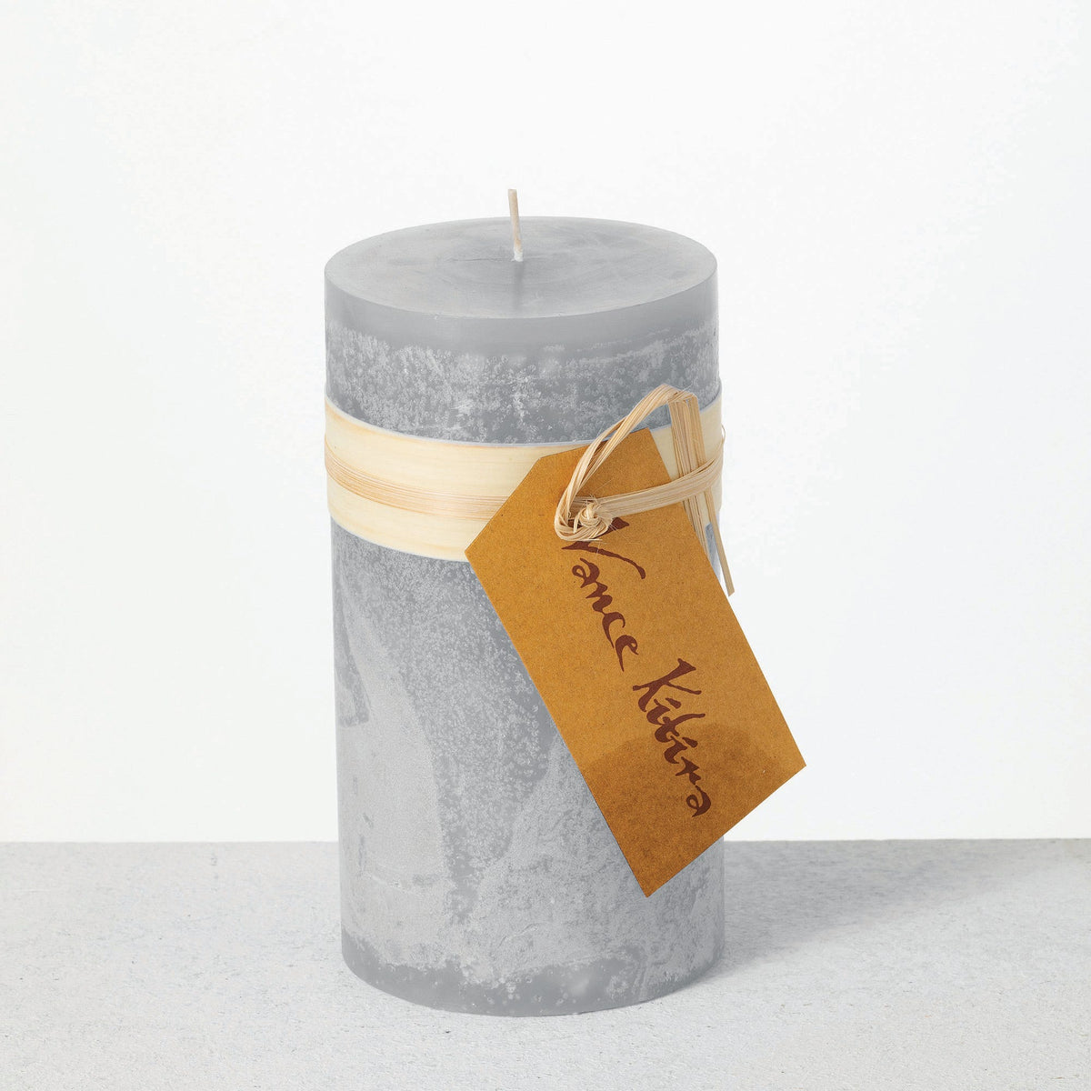 Timber Pillar Candle 3.25 x 6 Inch, A tall reddish brown pillar candle with a textured surface stands tied with a paper band and a hang tag.