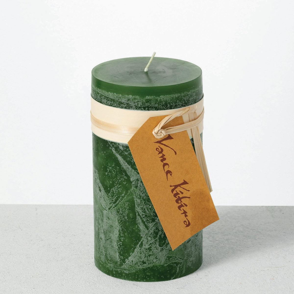 Timber Pillar Candle 3.25 x 6 Inch, A tall reddish brown pillar candle with a textured surface stands tied with a paper band and a hang tag.