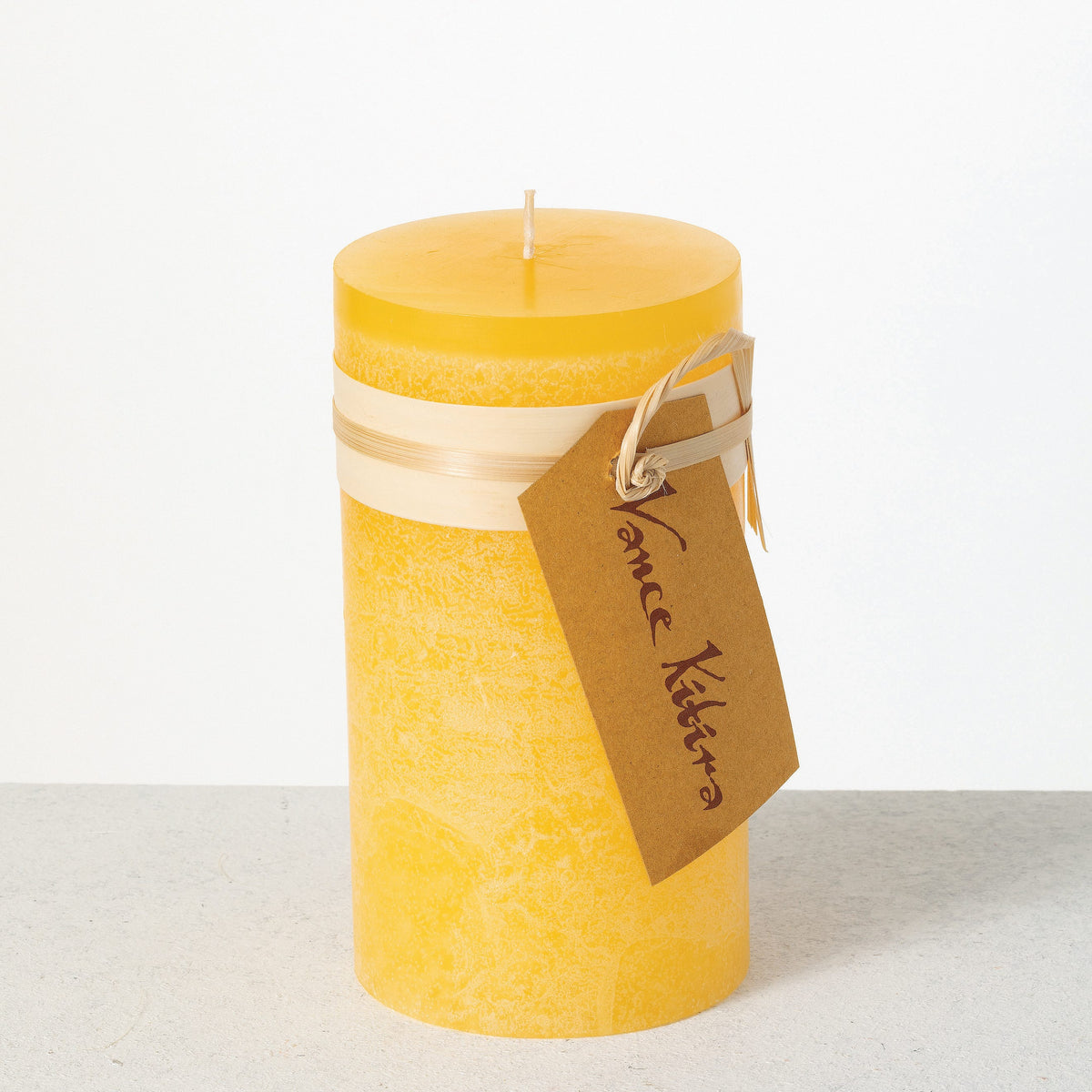 Timber Pillar Candle 3.25 x 6 Inch, A tall reddish brown pillar candle with a textured surface stands tied with a paper band and a hang tag.