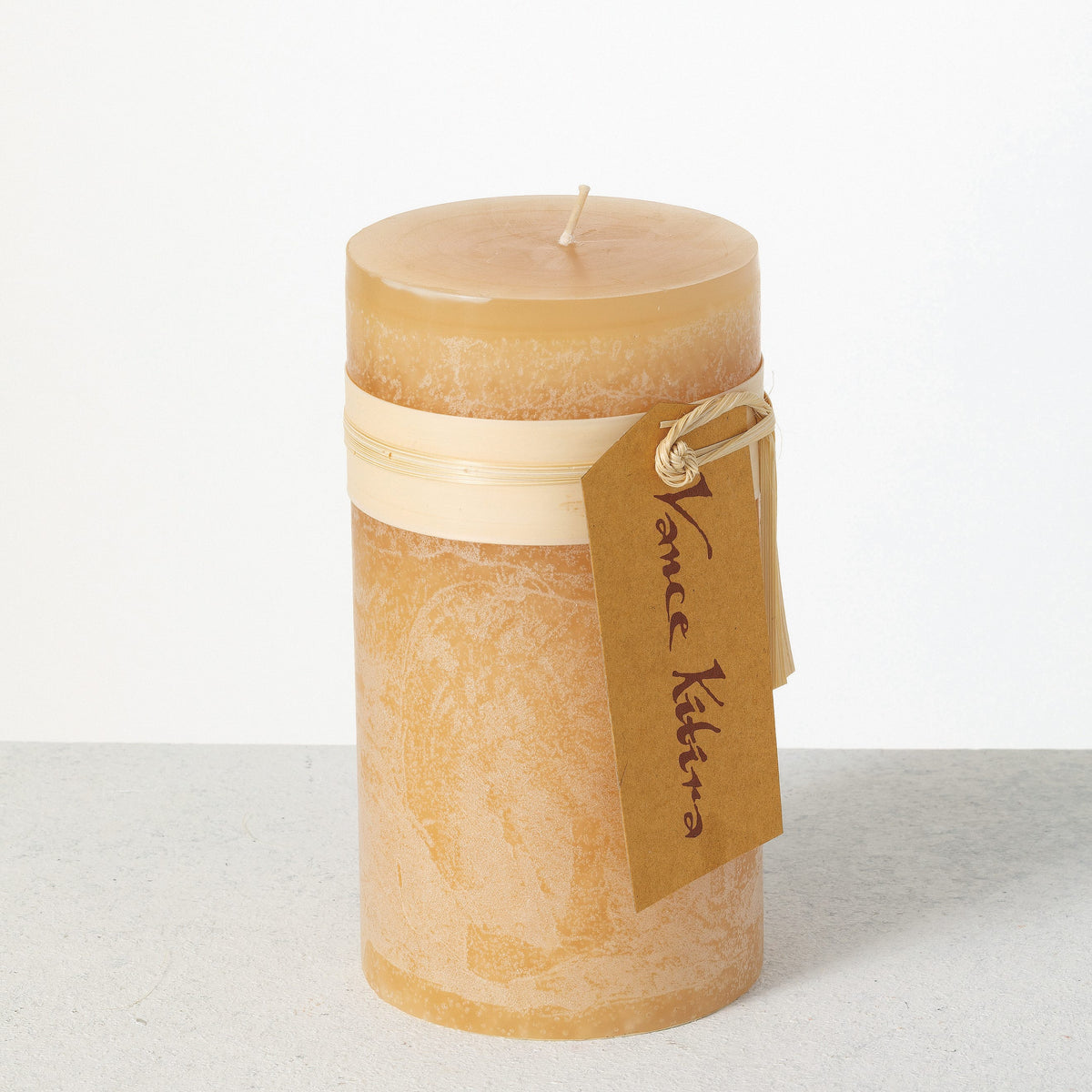 Timber Pillar Candle 3.25 x 6 Inch, A tall reddish brown pillar candle with a textured surface stands tied with a paper band and a hang tag.
