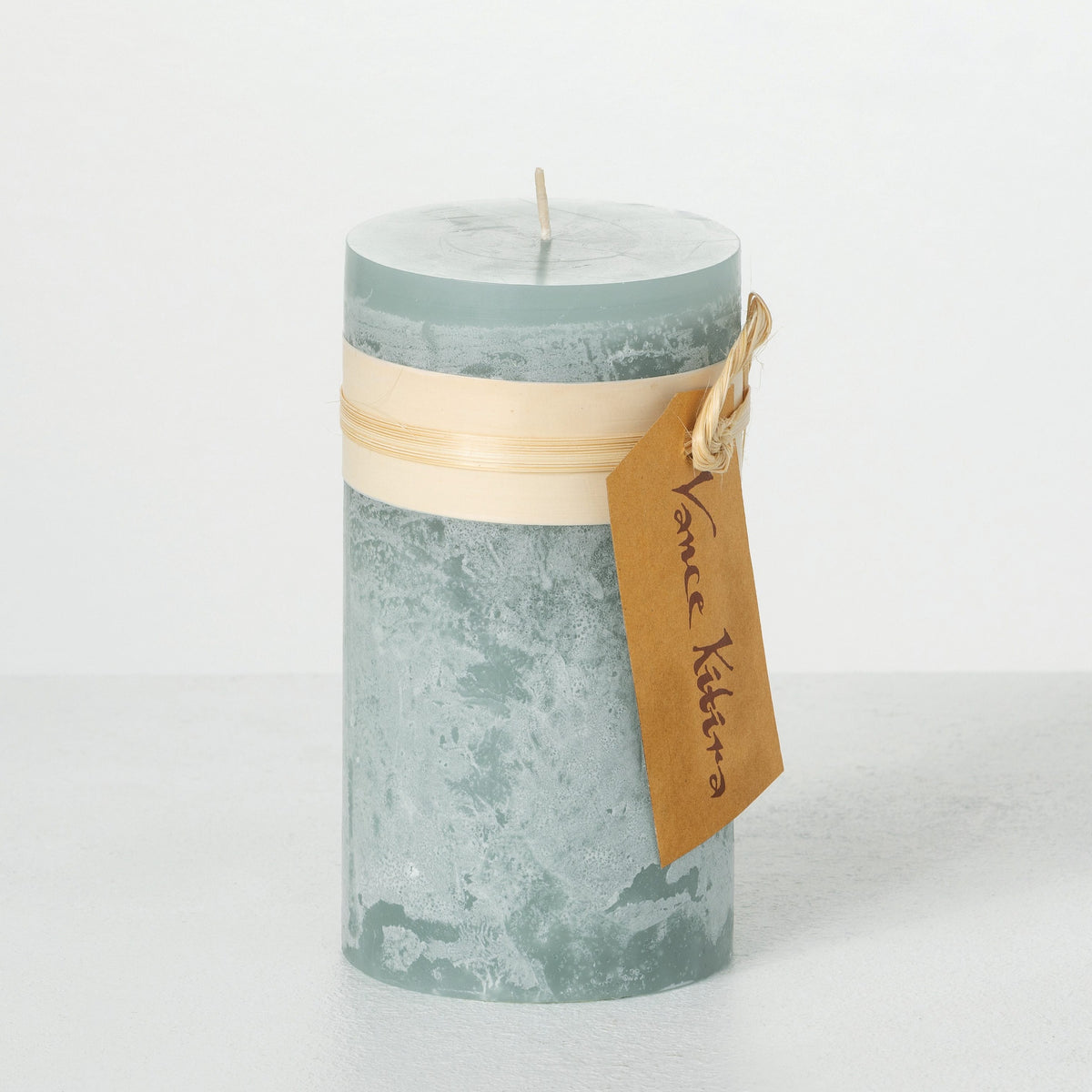 Timber Pillar Candle 3.25 x 6 Inch, A tall reddish brown pillar candle with a textured surface stands tied with a paper band and a hang tag.