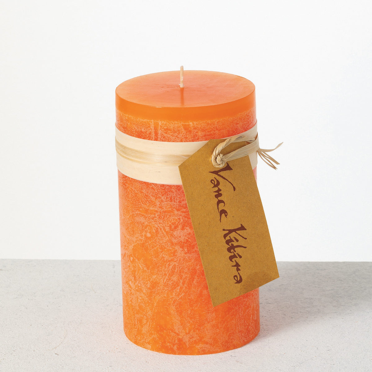 Timber Pillar Candle 3.25 x 6 Inch, A tall reddish brown pillar candle with a textured surface stands tied with a paper band and a hang tag.