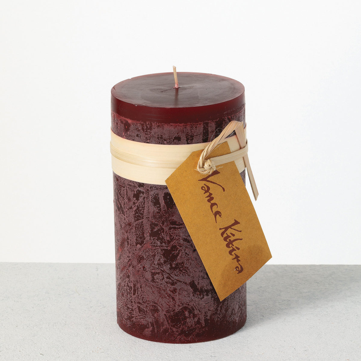 Timber Pillar Candle 3.25 x 6 Inch, A tall reddish brown pillar candle with a textured surface stands tied with a paper band and a hang tag.